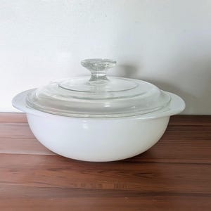 May include: A round, white milk glass casserole dish with integrated side handles and a matching clear glass lid featuring a central knob handle. The dish has a smooth, opaque finish and a clean, simple design suitable for serving or food storage.