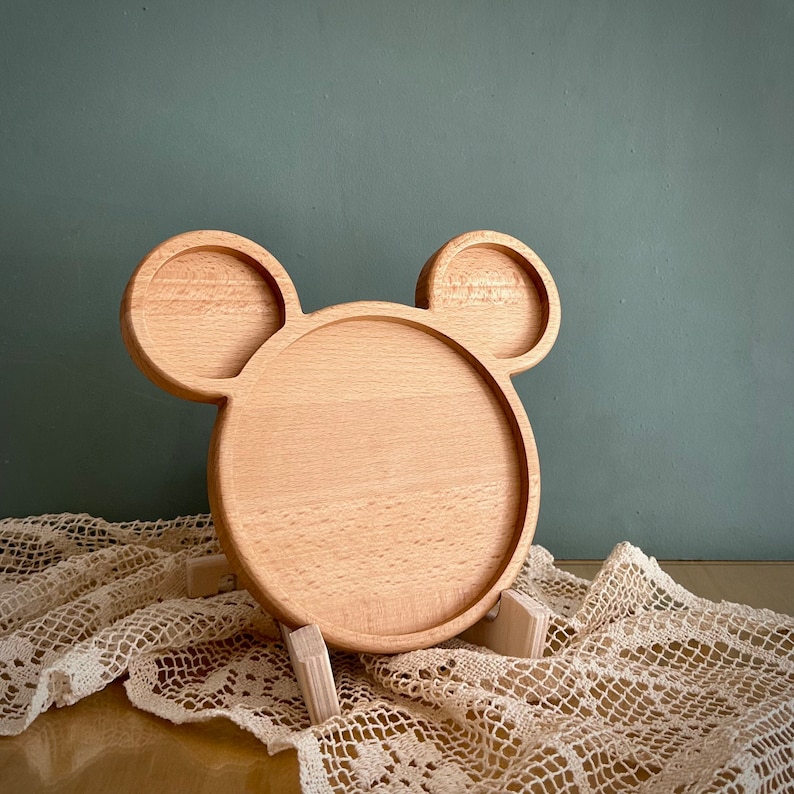 May include: A light-colored wooden serving tray in the shape of Mickey Mouse's head. The tray has a circular center and two smaller circular ears. It is supported by small wooden legs and sits on a white lace cloth.