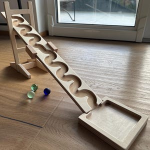 Puede incluir: Un juguete de circuito de canicas de madera con una pista ondulada y una estructura de soporte. La pista es de color marrón claro con un diseño tallado, y tres canicas de vidrio verdes y azules están cerca. El juguete está sobre un suelo de madera.