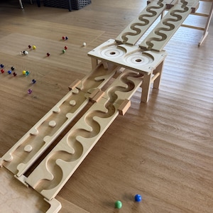 Op de afbeelding: Houten knikkerbaan met meerdere banen en niveaus. De natuurlijke houtkleurige structuur heeft gebogen en rechte paden waar knikkers doorheen kunnen rollen. Gekleurde knikkers in rood, groen, blauw, geel en paars liggen rond het speelgoed.