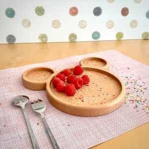 May include: A wooden plate in the shape of Mickey Mouse's head, holding fresh red raspberries. Two stainless steel serving utensils are on the left. The plate sits on a pink placemat, with colorful sprinkles scattered around.