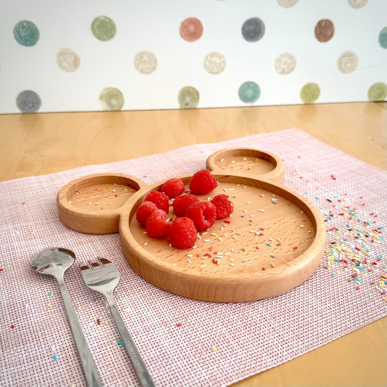 May include: A wooden plate in the shape of Mickey Mouse's head, holding fresh red raspberries. Two stainless steel serving utensils are on the left. The plate sits on a pink placemat, with colorful sprinkles scattered around.
