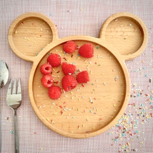 May include: A wooden plate in the shape of Mickey Mouse's head, with two ear-shaped sections. The plate holds fresh red raspberries and colorful sprinkles. A fork and spoon are visible on the left side, suggesting a meal setting.