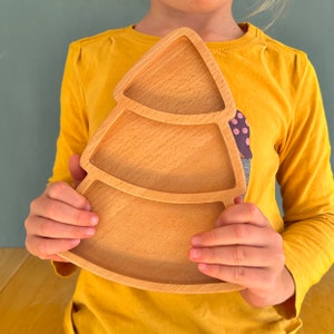 Op de afbeelding: Een houten, boomvormige serveerschaal met drie compartimenten. De schaal is gemaakt van lichtgekleurd hout en heeft een natuurlijke afwerking. De schaal wordt vastgehouden door een persoon die een gele trui draagt.