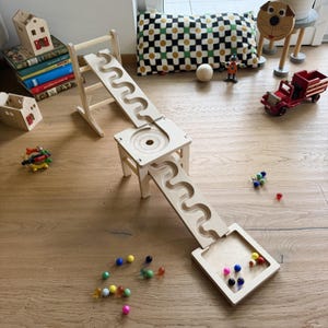 Könnte beinhalten: Eine Kugelbahn aus hellem Holz mit zwei langen, gewellten Bahnen, die durch eine quadratische Mittelplattform mit kreisförmiger Rille verbunden sind und in einer flachen quadratischen Basis enden, begleitet von mehreren kleinen, bunten Glaskugeln.