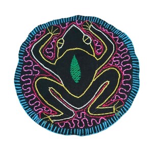 May include: A round, black embroidered patch featuring a stylized frog design. The frog is outlined in yellow and white thread, with a green leaf-shaped detail. The patch is bordered with blue, pink, and white stitching.