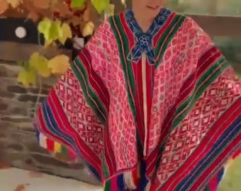 Könnte beinhalten: Ein farbenfrohes, gemustertes Poncho in Rosa, Rot, Grün und Blau. Der Poncho hat geometrische Designs und gestreifte Akzente mit farbenfrohen Fransen an den Kanten. Das Kleidungsstück ist Teil einer traditionellen Bekleidungskollektion.