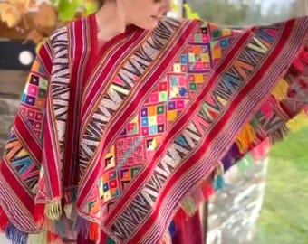 Könnte beinhalten: Ein lebendiger, handgewebter Poncho mit roter Basis, verziert mit bunten geometrischen Mustern und Fransen. Der Poncho weist eine Mischung aus leuchtenden Farben wie Rot, Gelb, Blau und Grün auf. Das Kleidungsstück ist Teil eines traditionellen Kleidungsensembles.