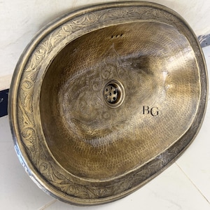 May include: Oval-shaped, hammered copper sink with an ornate floral border and interior design. The sink has a central drain and three faucet holes. The initials "BG" are etched on the right side of the sink.