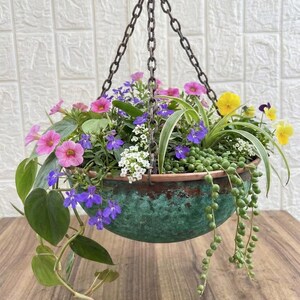 May include: A round, hammered copper-toned hanging planter with a weathered green patina finish, suspended by three dark metal chains. The planter is filled with a variety of colourful flowers, including pink, purple, yellow, and white blossoms.