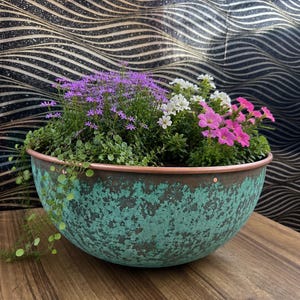 May include: A round, bowl-shaped planter with a weathered, teal-green patina finish and a smooth copper-coloured rim. The planter is filled with a dense arrangement of small purple flowers, white blossoms, and vibrant pink flowers with green foliage.