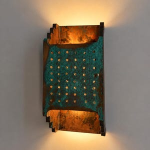 May include: A rectangular wall sconce featuring a weathered copper finish with a distinct teal-green patina. The front panel is perforated with a grid of small circular holes, allowing warm light to emit from the top and bottom openings.