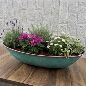 May include: An oval-shaped planter with a weathered teal patina finish and a copper-toned rim. The planter is filled with a variety of plants, including purple lavender, vibrant pink star-shaped flowers, green rosemary, white daisies, and trailing ivy.
