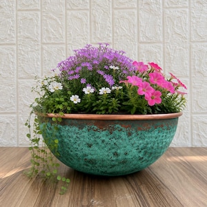 May include: A round, hammered copper bowl with a weathered, teal-green patina finish. The bowl is filled with a vibrant arrangement of small purple flowers, bright pink petunias, tiny white blossoms, and trailing green foliage.