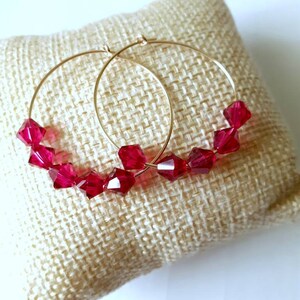 Peut inclure: Boucles d'oreilles créoles dorées avec des perles de cristal rouges. Les boucles d'oreilles sont sur une surface en tissu beige.