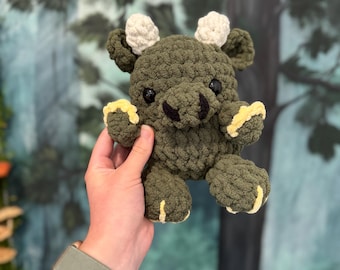 May include: A small, olive green crocheted plush creature with a rounded body, two white textured horns, black safety eyes, and yellow accents on the feet and hands. The plush has a thick, yarn-based texture and a compact, stuffed appearance.
