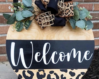 May include: Round wooden welcome sign with a black and leopard print design. The sign features the word "Welcome" in white script across a black stripe. It is decorated with a black and leopard print bow and greenery.