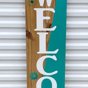 May include: A wooden welcome sign with the word "WELCOME" in large white letters. The sign has a teal painted section and is decorated with a bow and greenery at the top. The background is a white, horizontally-paneled surface.