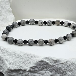 Peut inclure: Un bracelet de perles grises et noires. Le bracelet alterne de grosses perles grises rondes avec un motif marbré et de petites perles noires facettées. Le bracelet est présenté sur une surface blanche texturée.