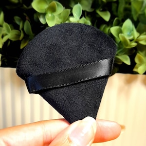 May include: A black velvet ring box with a black satin ribbon. The box is shaped like a triangle and is held in a hand.