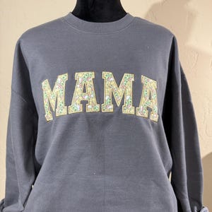May include: Gray sweatshirt with the word "MAMA" printed in large, yellow letters. The letters are filled with a pattern of small green and yellow images.