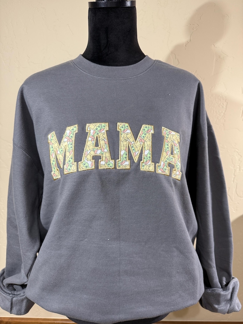 May include: Gray sweatshirt with the word "MAMA" printed in large, yellow letters. The letters are filled with a pattern of small green and yellow images.