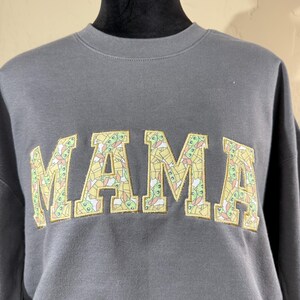 May include: A gray sweatshirt with the word "MAMA" in large, yellow letters. The letters are filled with a pattern of small, green, and yellow cartoon characters.