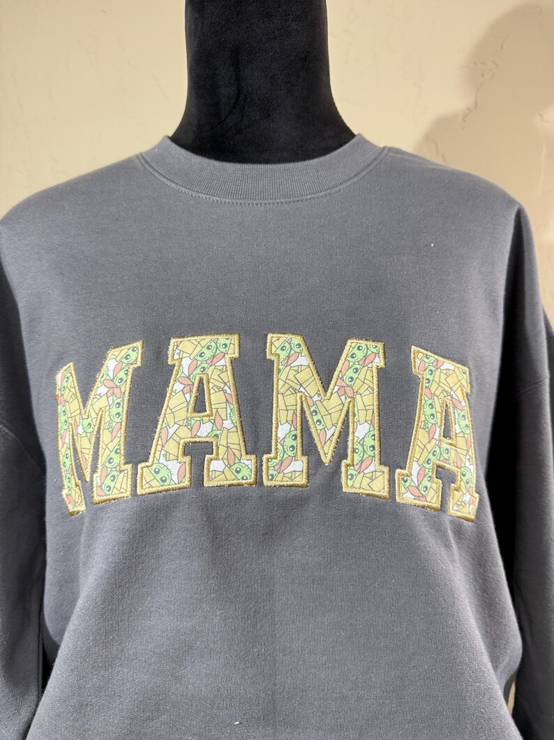 May include: A gray sweatshirt with the word "MAMA" in large, yellow letters. The letters are filled with a pattern of small, green, and yellow cartoon characters.