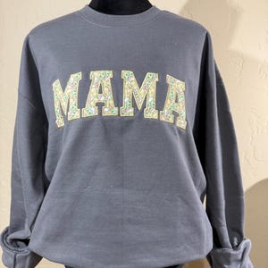 May include: A gray sweatshirt with the word "MAMA" printed on the front in yellow letters. The letters are filled with a pattern of small green and yellow images.
