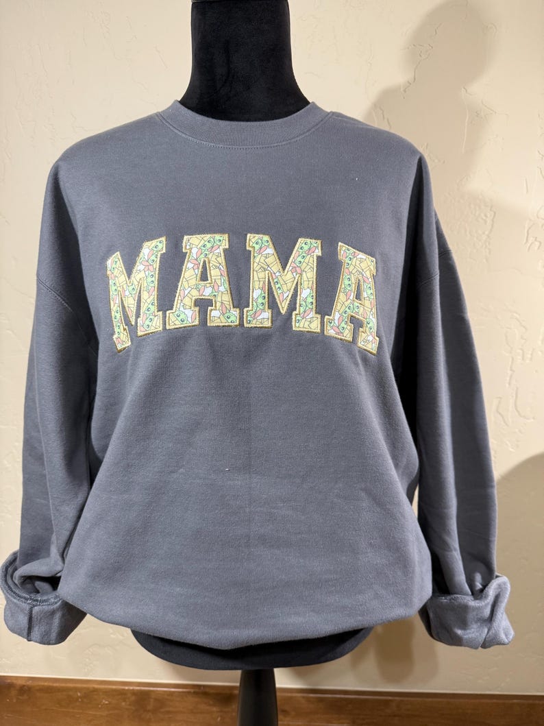 May include: A gray sweatshirt with the word "MAMA" printed on the front in yellow letters. The letters are filled with a pattern of small green and yellow images.