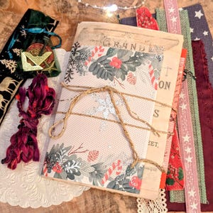 Könnte beinhalten: Eine Sammlung von Bastelmaterialien, darunter ein mit Bindfaden gebundenes Journal, dekorative Bänder und ein Samtbeutel. Das Journal hat ein florales Design mit dem Wort "GRAND". Rote, grüne und blau-sternförmige Bänder sind zu sehen.