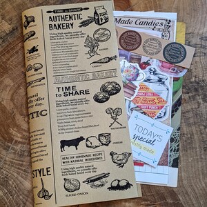 May include: A brown paper notebook with the words "Authentic Bakery" and "Time to Share" printed on the cover. The notebook is decorated with stickers and drawings of food, including vegetables, fruit, and bread. The notebook also has a sticker that says "Today's Special" and "Freshly Made".