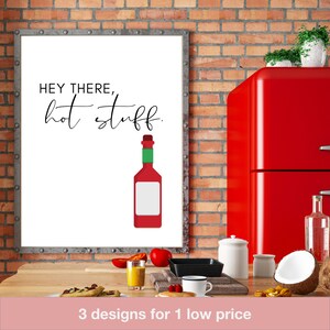 Puede incluir: Un cuadro enmarcado con el texto "HEY THERE, hot stuff" y una ilustración de una botella de salsa picante roja. El cuadro se muestra en una cocina con un refrigerador rojo y varios utensilios de cocina.