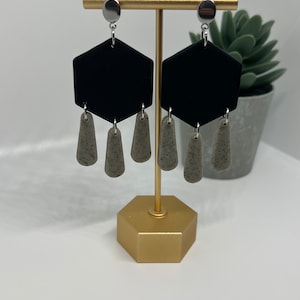 May include: A pair of black hexagon-shaped earrings with three dangling gray teardrop-shaped accents. The earrings are displayed on a gold-colored stand with a silver top. A small potted plant is in the background.