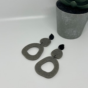 May include: A pair of gray statement earrings with a speckled texture. Each earring features two large, irregular oval shapes with a smaller circle in the center. The earrings have black accents.