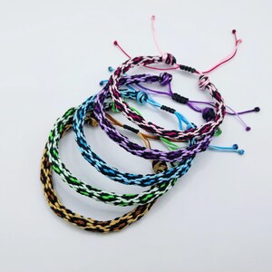 May include: A selection of woven bracelets in various colours. Each bracelet displays a distinct pattern and colour combination, including brown, green, blue, purple, and pink. The bracelets are adjustable with a knotted fastening.