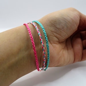 May include: Three braided bracelets in pink, teal, and a mix of pink and white are shown on a wrist. The bracelets are made of woven material, and the colours are vibrant. The bracelets are a type of jewellery.