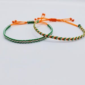 May include: Two friendship bracelets with a green, white and orange colour scheme. The bracelets are made of braided string and have a chevron pattern.