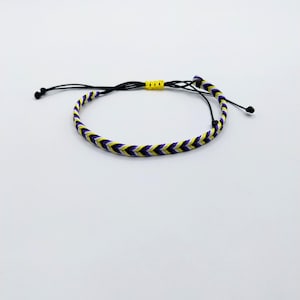 May include: A braided cord bracelet featuring a chevron pattern in yellow, white, black, and purple. The bracelet includes a black adjustable sliding cord closure with a small yellow accent bead and black knotted ends on each side of the closure.