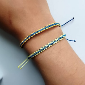 May include: Two friendship bracelets, one blue and yellow, the other blue and green, are worn on a person's wrist.