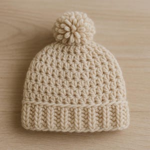 May include: A cream-coloured, crocheted beanie hat with a ribbed brim and a large pom-pom on top. The hat is made of a soft, textured yarn and is suitable for cold weather. The hat is a neutral colour and can be worn by anyone.