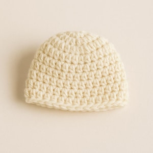 Puede incluir: Un pequeño gorro de ganchillo de color crema. El gorro está hecho con un patrón de puntadas apretadas, lo que le da una apariencia texturizada. El gorro es probablemente para un bebé o un niño pequeño. El gorro es de color liso y tiene un diseño simple y clásico.
