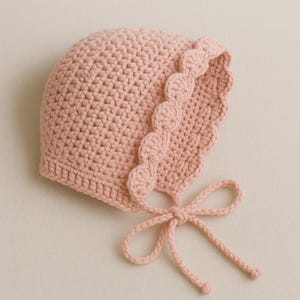 Puede incluir: Un gorro de bebé de ganchillo rosa claro con borde festoneado y cierre de lazo. El gorro está hecho de hilo suave y está diseñado para la cabeza de un bebé. Artículo hecho a mano.