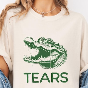 May include: A white t-shirt featuring a green alligator graphic and the text "TEARS" printed in green below the graphic. The t-shirt is modelled on a person wearing a denim jacket.