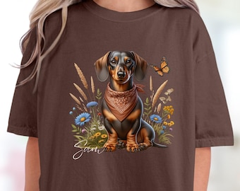 Op de afbeelding: Espresso-kleurig T-shirt met een illustratie van een teckel. De hond draagt een bandana en zit in een bloemenveld met een vlinder. Het woord "Sam" staat in cursief onder de hond.