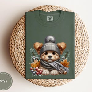 Op de afbeelding: Een groen T-shirt met een schattige illustratie van een hond die een grijze gebreide muts en sjaal draagt. De hond is omringd door herfstbladeren en bessen. De tekst "Sam" is op het T-shirt gedrukt.