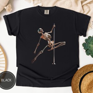 May include: Black t-shirt featuring a skeleton figure pole dancing. The skeleton is brown and white, with a blue cap. The pole is silver. The word "BLACK" is printed in a black circle at the bottom left.