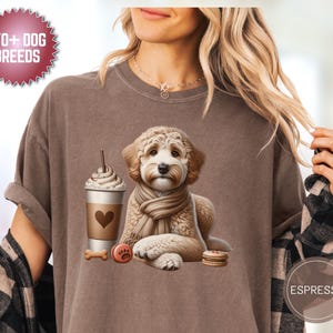 Op de afbeelding: Bruin T-shirt met een afbeelding van een hond met een sjaal, een koffiekopje, een bot en een koekje. Het shirt heeft de tekst "170+ DOG BREEDS" op een cirkelvormig embleem. Het woord "Espresso" staat op een cirkel.