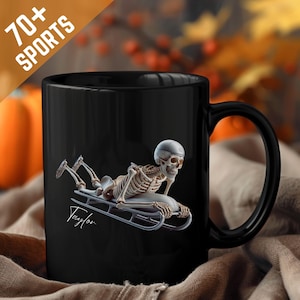Puede incluir: Taza de cerámica negra con un esqueleto con casco en un trineo. El esqueleto está en posición de carrera. La taza tiene el texto "70+ SPORTS" y el nombre "Taylor".