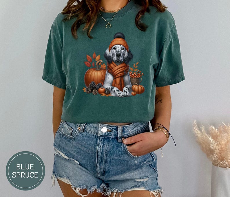 May include: A teal blue t-shirt with a white and brown dog wearing a brown and orange scarf and a brown and white hat. The dog is surrounded by fall leaves, pumpkins, and acorns.
